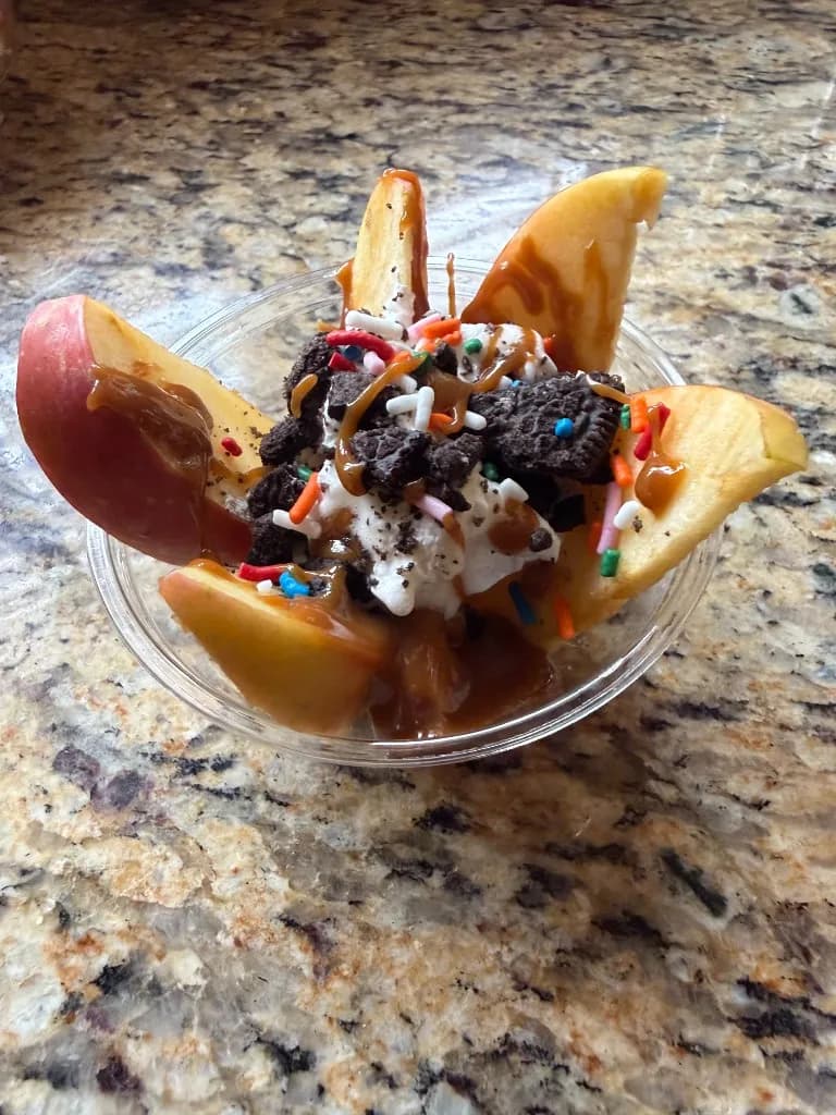 Apple slices with caramel, whipped cream, crushed chocolate cookies and rainbow sprinkles